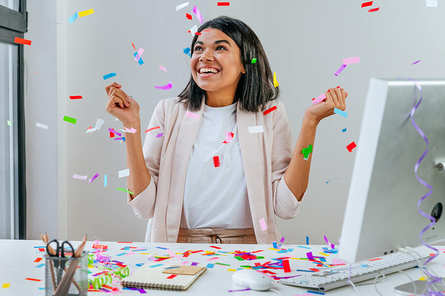 A woman joyfully celebrates at her desk, surrounded by confetti, commemorating work anniversaries with personalized chocolate gifts