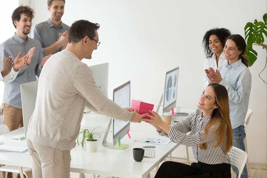 A woman at a desk receiving applause from a group of people. Corporate gifting theme, an employee receiving a gift