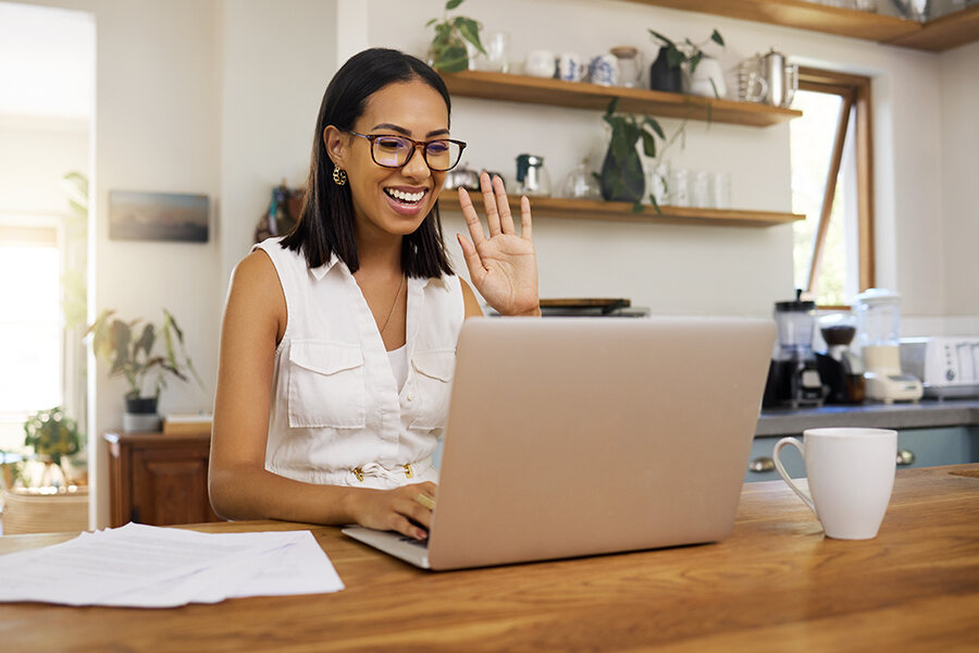 A smiling woman in glasses sits at a table with a laptop, working remotely