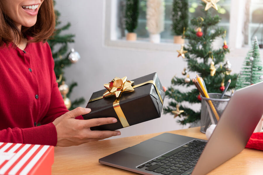 A woman holds a gift box in one hand and a laptop in the other, symbolizing corporate gifting solution